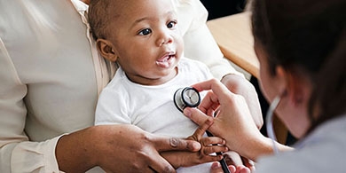 Photo of smiling baby with a stethoscope on chest being held by a physician during an exam.